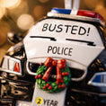 Load image into Gallery viewer, Personalized police car Christmas ornament featuring a black and white cop car design with BUSTED text, police lights and festive wreath detail. Customized with a name and year on the front banner. Glossy resin finish with bold black and white colors creates a fun and playful holiday look. Mostly 2D ornament with raised 3D details on the front only, lightweight and perfect for Christmas tree decor and police themed gifts.
