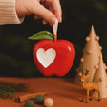 Load image into Gallery viewer, Red apple-shaped container with a heart cutout, held by a hand, on a dark background with decorative elements.
