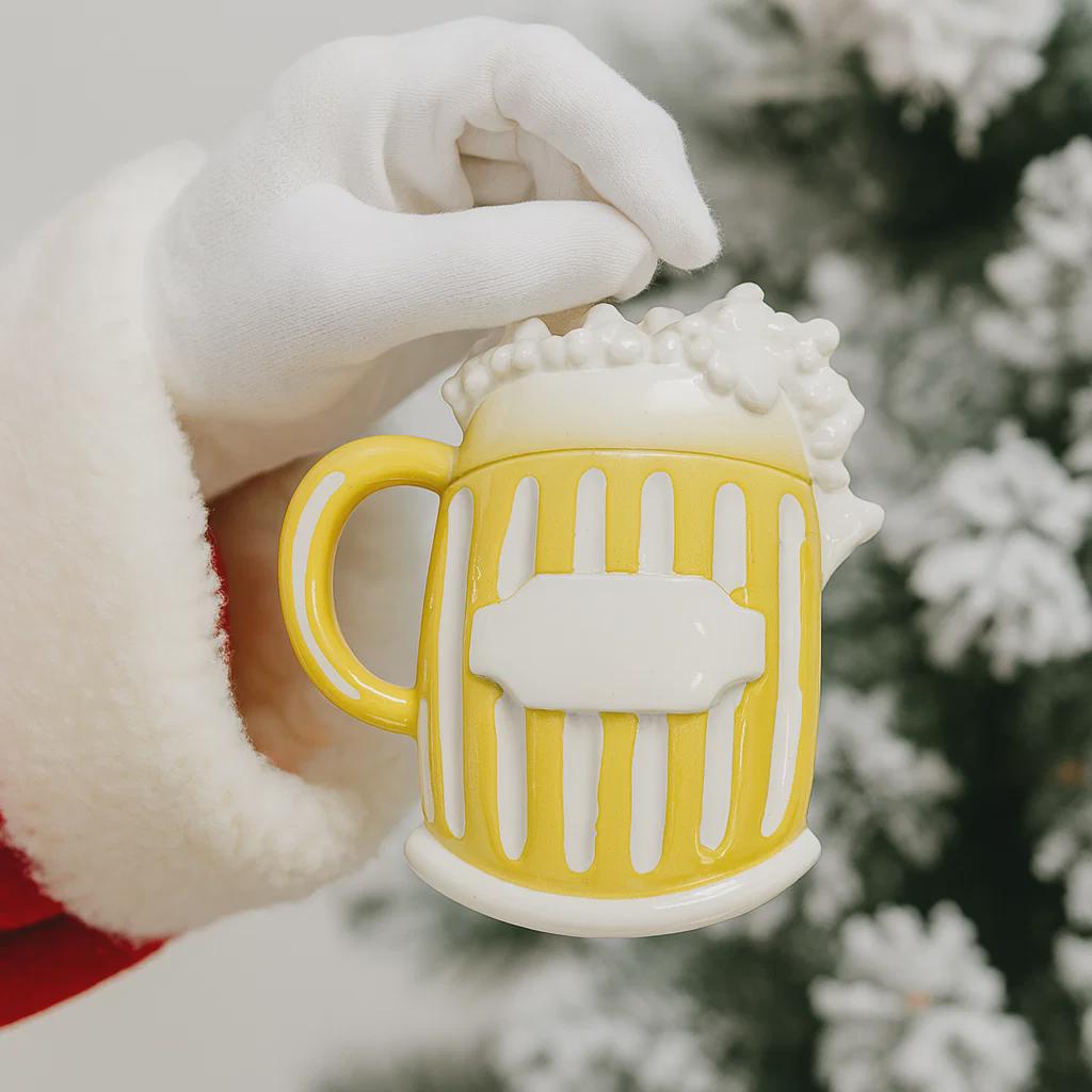 Yellow mug ornament held by a gloved hand with a blurred Christmas tree background