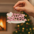 Load image into Gallery viewer, A personalized Little Sister ornament featuring soft pink layered lettering with heart accents and floral details. Designed in gentle pastel tones with a smooth resin finish and subtle glitter highlights. The ornament includes handwritten personalization with a baby name and year, created as a mostly 2D ornament with raised 3D details on the front only. Lightweight and easy to hang, perfect for celebrating a new baby sister and her first Christmas.
