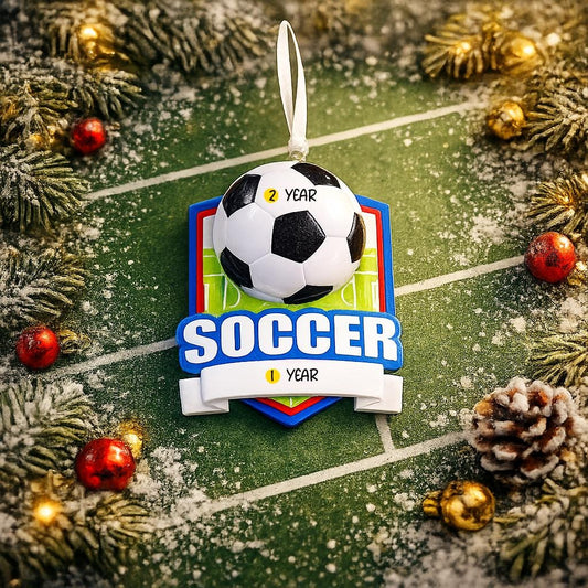 Personalized soccer Christmas ornament featuring a classic black and white soccer ball on a shield style background, customizable name area on the front banner and customizable year area on the soccer ball, glossy finish with raised 3D front details and flat 2D back, white hanging ribbon, displayed on a snowy soccer field themed holiday background with pine branches pinecones and festive decorations.