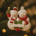 Load image into Gallery viewer, Decorative Christmas ornament of two snowmen holding a wreath and small tree against a blurred festive background.
