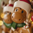 Load image into Gallery viewer, Close-up of a reindeer figurine wearing a Santa hat and green scarf with a blurred background.
