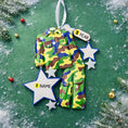 Load image into Gallery viewer, A glossy resin Army soldier uniform ornament in green brown and blue camouflage colors features a military jacket and pants shape with raised buttons and pockets, white and blue star accents, light snow texture, a smooth painted cartoon style finish, and handwritten personalization showing a name and year on the attached tag, designed as a mostly 2D ornament with raised 3D details on the front only for Christmas tree decor.

