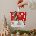 Load image into Gallery viewer, Decorative ornament with footballs, helmets, and stars on a red background, held by a hand.
