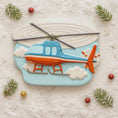 Load image into Gallery viewer, Decorative cookie shaped like a helicopter on a snowy background with pine branches and berries.
