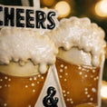 Load image into Gallery viewer, Personalized Cheers and Beers Christmas ornament featuring a raised 3D front with two frothy beer mugs clinking together. The design includes bold “Cheers” and “Beers” lettering, detailed foam texture, and warm amber beer tones. The ornament has a flat 2D back for easy hanging. Hand-added personalization allows two names and a year. Styled on a rustic wooden background with Christmas lights, greenery, and festive decor for a cozy holiday atmosphere.

