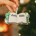Load image into Gallery viewer, Personalized Holy Bible Christmas Ornament featuring an open scripture design with elegant “Holy Bible” lettering across the pages. The left page is customized with a name and the right page displays the year, creating a meaningful faith based keepsake. Surrounded by soft greenery and delicate flowers, this religious ornament is perfect for baptisms, baby dedications, first communions, or celebrating a Christ centered Christmas. A thoughtful spiritual gift for families who cherish their faith.

