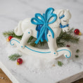 Load image into Gallery viewer, Decorative rocking horse with a blue bow on a snowy surface with Christmas decorations.
