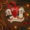 Load image into Gallery viewer, Decorative Christmas ornament with penguins and a heart, surrounded by festive items on a wooden surface.
