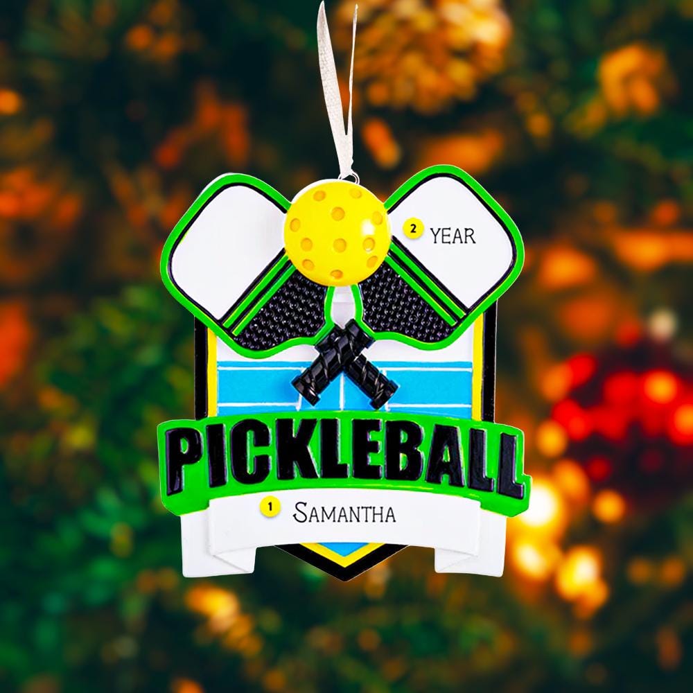 Personalized pickleball Christmas ornament featuring crossed pickleball paddles and a yellow pickleball on a shield style background, customizable name area on the front banner and customizable year area on the ball, glossy finish with raised 3D front details and flat 2D back, white hanging ribbon, displayed on a snowy pickleball court themed holiday background with pine branches and festive decorations.