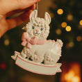Load image into Gallery viewer, A glossy resin baby girl llama ornament features a white smiling llama with soft pink accents, raised textured fur details, pink bow and scarf elements, glitter highlights, and a smooth painted cartoon style finish, styled with warm holiday lights and nursery themed props, handwritten personalization showing the baby name on the front banner and the year placed on the llama body, designed as a mostly 2D ornament with raised 3D details on the front only.
