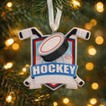 Load image into Gallery viewer, Personalized hockey Christmas ornament featuring a hockey puck and crossed hockey sticks design, customizable name area on the front banner and customizable year area on the puck, glossy finish with raised 3D front details and flat 2D back, white hanging ribbon, displayed on a snowy ice rink themed holiday background with hockey accessories and festive winter decorations.
