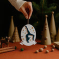 Load image into Gallery viewer, Personalized gymnastics Christmas ornament featuring an oval white design with a navy blue gymnast silhouette performing a handstand pose, surrounded by gold and blue star accents, customizable year displayed on the upper area, customizable name space on the bottom banner, smooth glossy finish with raised 3D front details and flat 2D back, white hanging ribbon, photographed on a soft pink snowy holiday background with winter greenery, festive ornaments, and warm seasonal styling.

