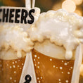 Load image into Gallery viewer, Personalized Cheers and Beers Christmas ornament featuring a raised 3D front with two frothy beer mugs clinking together. The design includes bold “Cheers” and “Beers” lettering, detailed foam texture, and warm amber beer tones. The ornament has a flat 2D back for easy hanging. Hand-added personalization allows two names and a year. Styled on a rustic wooden background with Christmas lights, greenery, and festive decor for a cozy holiday atmosphere.

