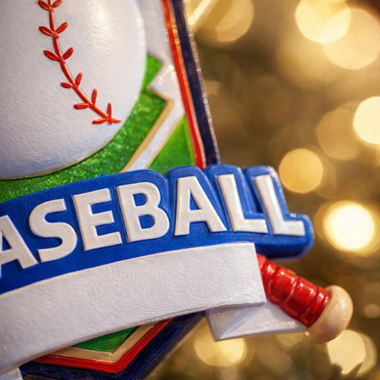 Personalized baseball Christmas ornament featuring a classic baseball and crossed bats design on a shield style background, customizable name area on the front banner and customizable year area on the baseball, glossy finish with raised 3D front details and flat 2D back, white hanging ribbon, displayed on a snowy baseball field themed holiday background with pine branches and festive decorations.