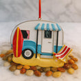 Load image into Gallery viewer, Decorative ornament of a beach-themed camper with a surfboard and chair on pebbles.
