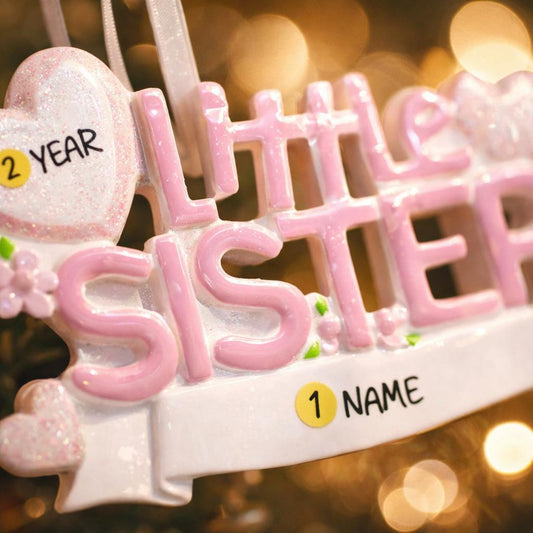 A personalized Little Sister ornament featuring soft pink layered lettering with heart accents and floral details. Designed in gentle pastel tones with a smooth resin finish and subtle glitter highlights. The ornament includes handwritten personalization with a baby name and year, created as a mostly 2D ornament with raised 3D details on the front only. Lightweight and easy to hang, perfect for celebrating a new baby sister and her first Christmas.