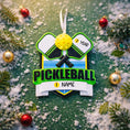 Load image into Gallery viewer, Personalized pickleball Christmas ornament featuring crossed pickleball paddles and a yellow pickleball on a shield style background, customizable name area on the front banner and customizable year area on the ball, glossy finish with raised 3D front details and flat 2D back, white hanging ribbon, displayed on a snowy pickleball court themed holiday background with pine branches and festive decorations.
