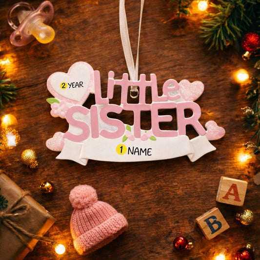 A personalized Little Sister ornament featuring soft pink layered lettering with heart accents and floral details. Designed in gentle pastel tones with a smooth resin finish and subtle glitter highlights. The ornament includes handwritten personalization with a baby name and year, created as a mostly 2D ornament with raised 3D details on the front only. Lightweight and easy to hang, perfect for celebrating a new baby sister and her first Christmas.