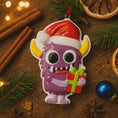 Load image into Gallery viewer, Christmas ornament with a purple monster holding a gift on a wooden surface with festive decorations.
