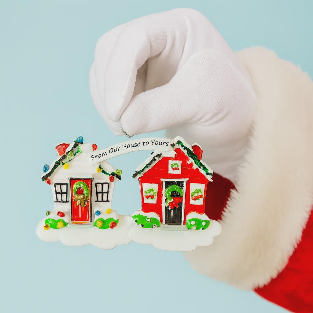 Christmas ornament shaped like a house with 'From Our House to Yours' text, held by a gloved hand.