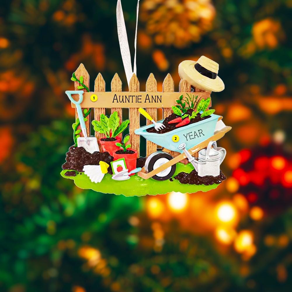 Personalized gardener ornament featuring a detailed garden scene with wooden fence wheelbarrow potted plants gardening tools and soil elements. The design includes a customizable name on the fence and a year on the base making it a thoughtful keepsake for garden lovers. The ornament has a warm rustic farmhouse style and is displayed with a red hanging ribbon suitable for Christmas trees seasonal decor and gift displays.