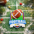 Load image into Gallery viewer, Personalized football Christmas ornament featuring a brown football with stitched details on a shield style background and goalpost design, customizable name area on the front banner and customizable year area on the football, glossy finish with raised 3D front details and flat 2D back, white hanging ribbon, displayed on a snowy football field themed holiday background with pine branches and festive decorations.
