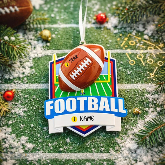 Personalized football Christmas ornament featuring a brown football with stitched details on a shield style background and goalpost design, customizable name area on the front banner and customizable year area on the football, glossy finish with raised 3D front details and flat 2D back, white hanging ribbon, displayed on a snowy football field themed holiday background with pine branches and festive decorations.