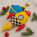 Load image into Gallery viewer, Colorful rocket-shaped Christmas ornament on a snowy background with pine branches and red ornaments.
