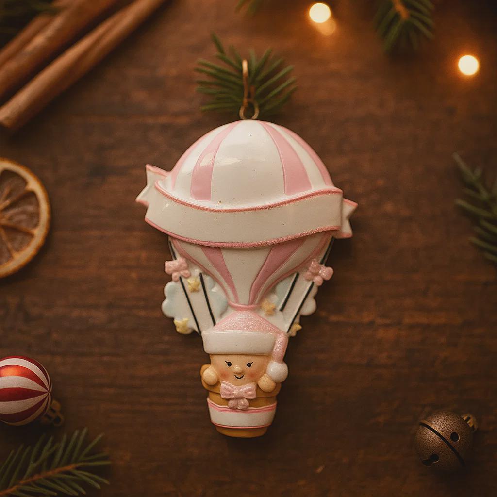 Personalized baby first Christmas ornament featuring a soft pastel pink hot air balloon design with a sweet baby character inside the basket. Handwritten baby name and year or Baby’s First Christmas text displayed on the balloon. Smooth resin finish with gentle glitter accents, nursery themed colors, lightweight structure, mostly 2D ornament with raised 3D details on the front only, designed for baby Christmas tree decor and keepsake gifting.