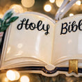 Load image into Gallery viewer, Personalized Holy Bible Christmas Ornament featuring an open scripture design with elegant “Holy Bible” lettering across the pages. The left page is customized with a name and the right page displays the year, creating a meaningful faith based keepsake. Surrounded by soft greenery and delicate flowers, this religious ornament is perfect for baptisms, baby dedications, first communions, or celebrating a Christ centered Christmas. A thoughtful spiritual gift for families who cherish their faith.
