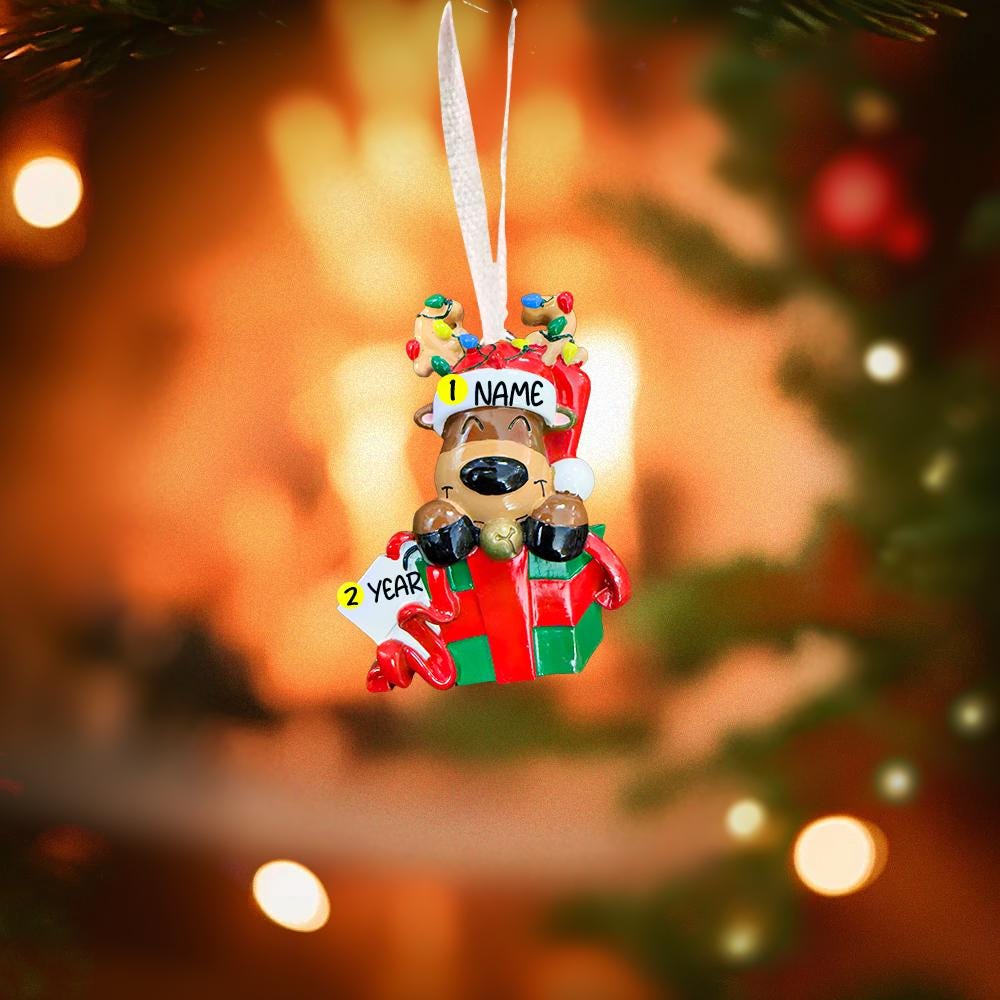Personalized Christmas ornament with a teddy bear on a gift box against a blurred Christmas tree background.