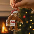 Load image into Gallery viewer, This playful Bigfoot Christmas ornament features a cheerful sasquatch character wearing a Santa hat and holding a gift, finished with a glossy surface that highlights the raised details and festive colors. The design brings a fun and whimsical holiday vibe, making it a standout decoration for Christmas trees, shelves, or seasonal displays, especially for Bigfoot lovers and collectors of unique holiday ornaments.
