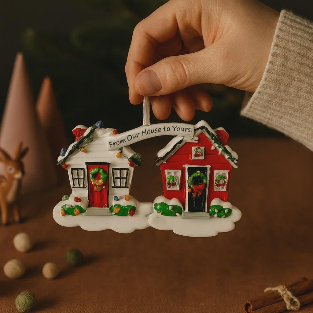 Hand holding a Christmas ornament shaped like two houses with a 'From Our House to Yours' sign.