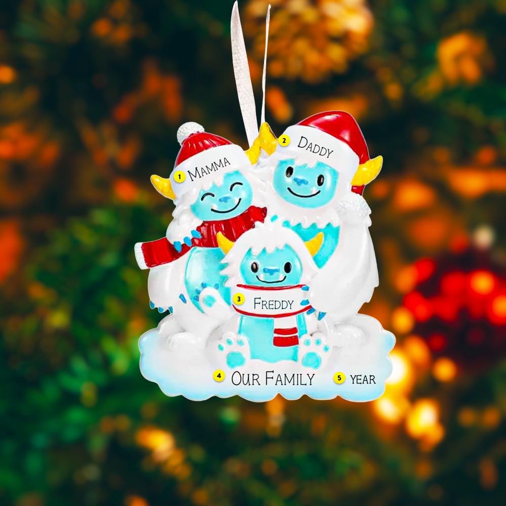 Christmas ornament with yeti family of three in winter clothing against a blurred Christmas tree background.