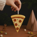 Load image into Gallery viewer, Pizza slice ornament held by a hand with Christmas decorations in the background
