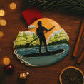 Load image into Gallery viewer, Decorative ornament of a person paddleboarding on water with trees and sunset, placed on a wooden surface.

