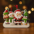 Load image into Gallery viewer, Christmas ornament with Santa Claus, snowman, elf, and penguin on a wooden surface with blurred lights in the background.
