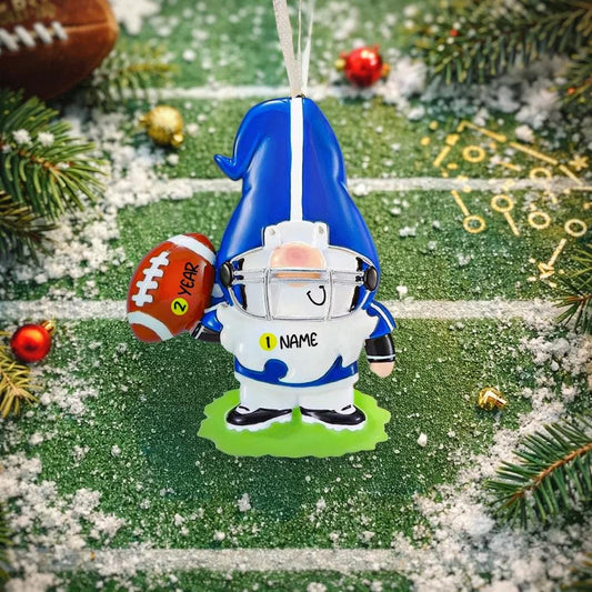 Personalized football gnome Christmas ornament featuring a blue and white gnome character wearing a football helmet and holding a football, customizable name area on the front body and customizable year area on the football, glossy finish with raised 3D front details and flat 2D back, white hanging ribbon, displayed on a snowy football field themed holiday background with pine branches and festive decorations.