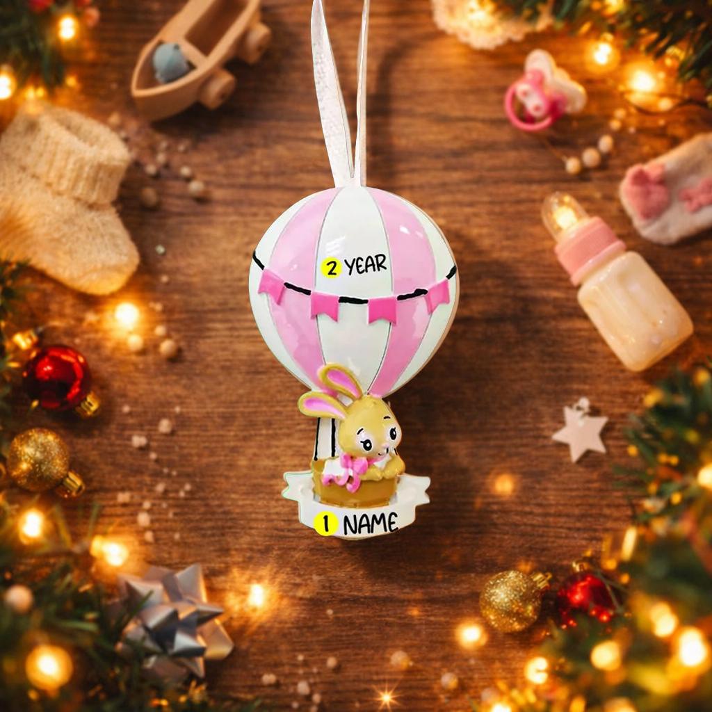 A personalized baby girl ornament featuring a cute bunny riding a pastel pink hot air balloon. Soft pink and white tones with delicate bow details create a sweet and cozy look. Handwritten personalization includes baby name and year. Mostly 2D ornament with raised 3D details on the front only, lightweight and easy to hang, perfect for baby first Christmas, nursery decor, and toddler keepsakes.