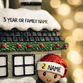Load image into Gallery viewer, Personalized Christmas house ornament featuring a cozy winter home decorated with snow, wreaths, garlands, and festive details. Two smiling characters wearing Santa hats peek from the front, each customizable with a name, while the roof displays a year or family name. Designed with a detailed 3D raised front and smooth finish, this ornament is perfect for Christmas tree decor, family keepsakes, new home celebrations, and holiday gifting.

