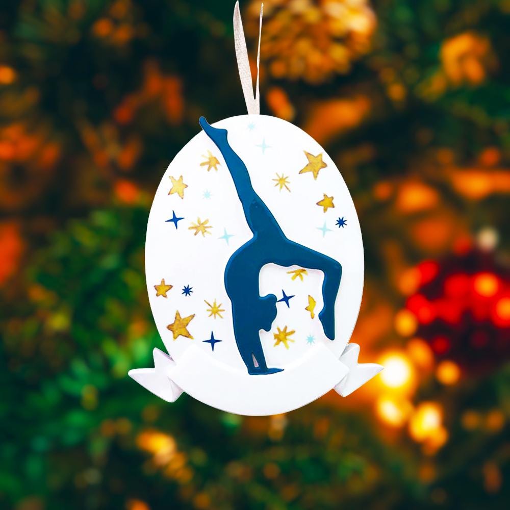 Personalized gymnastics Christmas ornament featuring an oval white design with a navy blue gymnast silhouette performing a handstand pose, surrounded by gold and blue star accents, customizable year displayed on the upper area, customizable name space on the bottom banner, smooth glossy finish with raised 3D front details and flat 2D back, white hanging ribbon, photographed on a soft pink snowy holiday background with winter greenery, festive ornaments, and warm seasonal styling.
