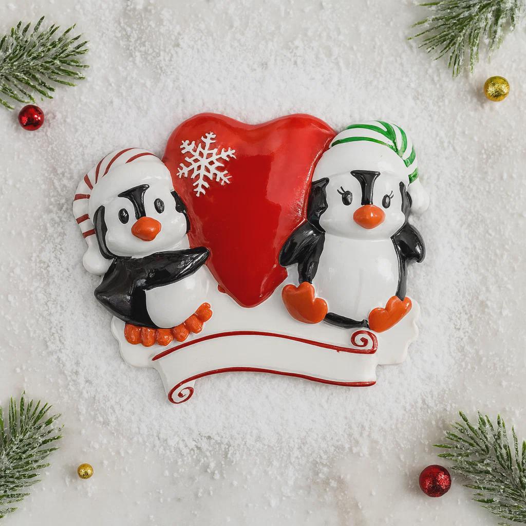 Decorative ornament with two penguins holding a red heart on a snowy background.