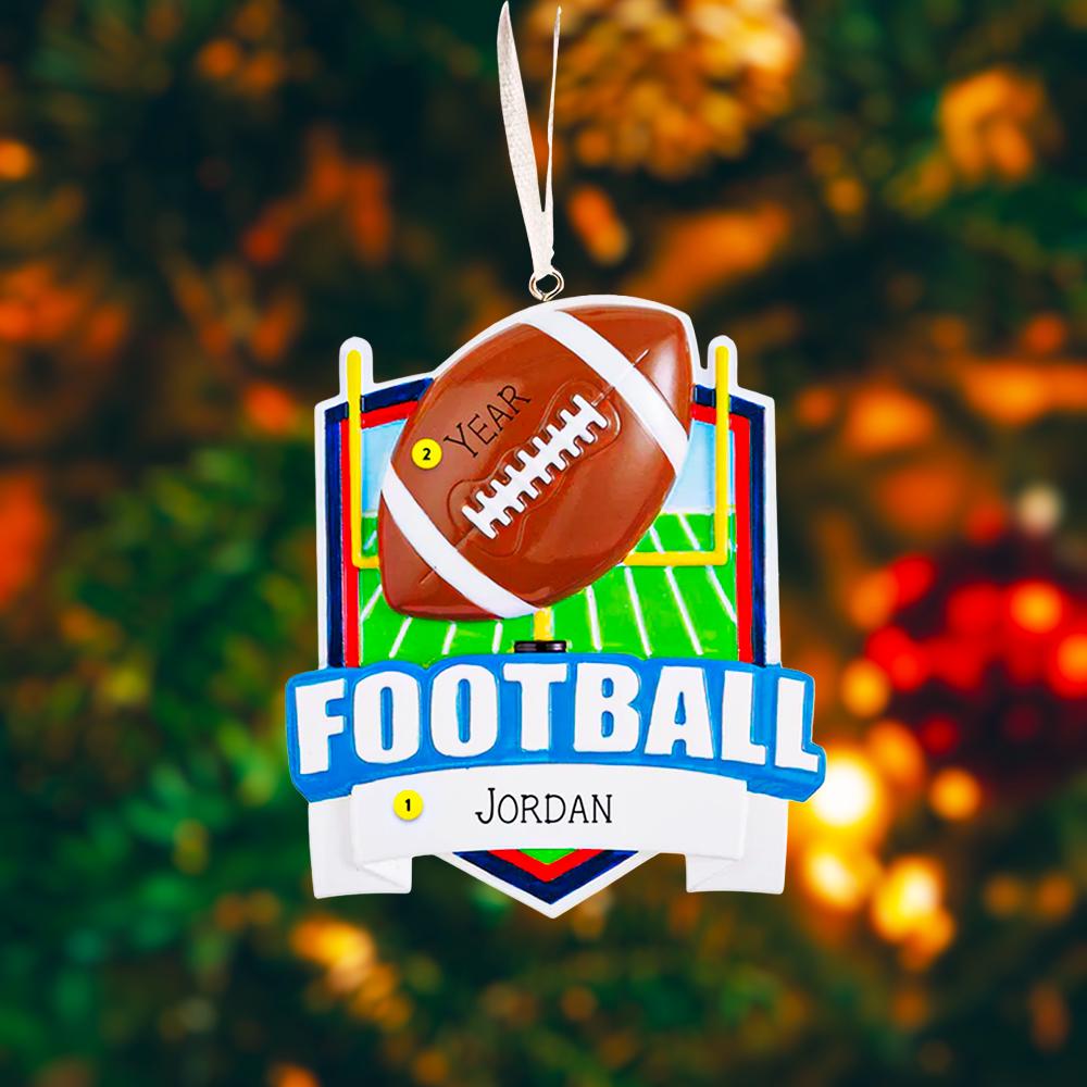 Personalized football Christmas ornament featuring a brown football with stitched details on a shield style background and goalpost design, customizable name area on the front banner and customizable year area on the football, glossy finish with raised 3D front details and flat 2D back, white hanging ribbon, displayed on a snowy football field themed holiday background with pine branches and festive decorations.