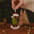 Load image into Gallery viewer, Decorative pickle ornament with a Santa hat and sunglasses, held by a hand against a festive background.
