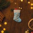 Load image into Gallery viewer, A glossy resin baby boy stocking ornament features a soft pastel blue Christmas stocking shape with a raised glitter style baby figure on the front, white cuff detail, small star accents, and smooth painted surfaces, surrounded by warm holiday lights and nursery themed props, handwritten personalization showing one baby name on the top cuff and the year written near the bottom of the stocking, designed as a mostly 2D ornament with raised 3D details on the front only.
