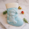 Load image into Gallery viewer, A glossy resin baby boy stocking ornament features a soft pastel blue Christmas stocking shape with a raised glitter style baby figure on the front, white cuff detail, small star accents, and smooth painted surfaces, surrounded by warm holiday lights and nursery themed props, handwritten personalization showing one baby name on the top cuff and the year written near the bottom of the stocking, designed as a mostly 2D ornament with raised 3D details on the front only.
