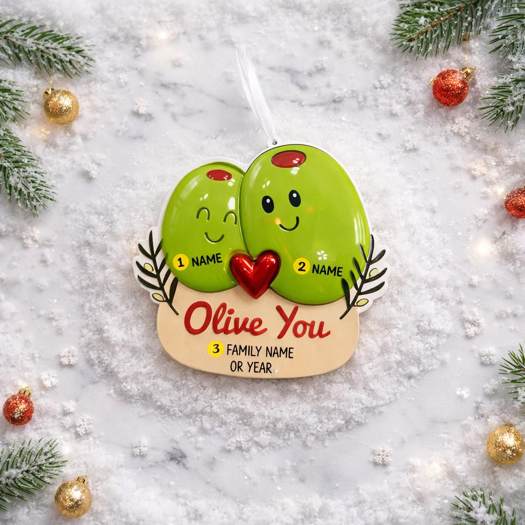 Decorative ornament with two smiling green olives holding a heart, surrounded by Christmas decorations on a snowy background.