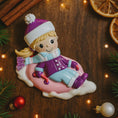 Load image into Gallery viewer, Christmas ornament of a girl on a sled with festive decorations around
